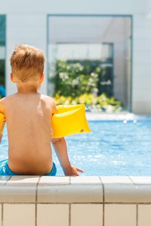Back view cute adorable little caucasian blond boy kid wear inflatable armband sit poolside indoor pool edge before swimming hotel spa thermal resort. Happy child enjoy summer vacation travel family.