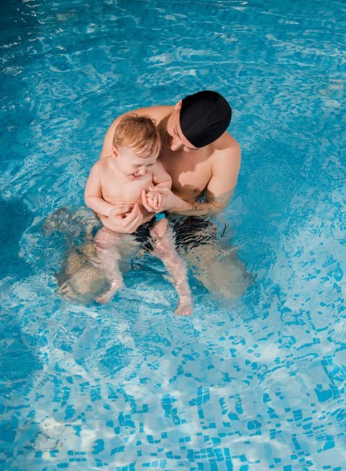 overhead view of smiling swim coach in swimming cap training toddler boy in swimming pool