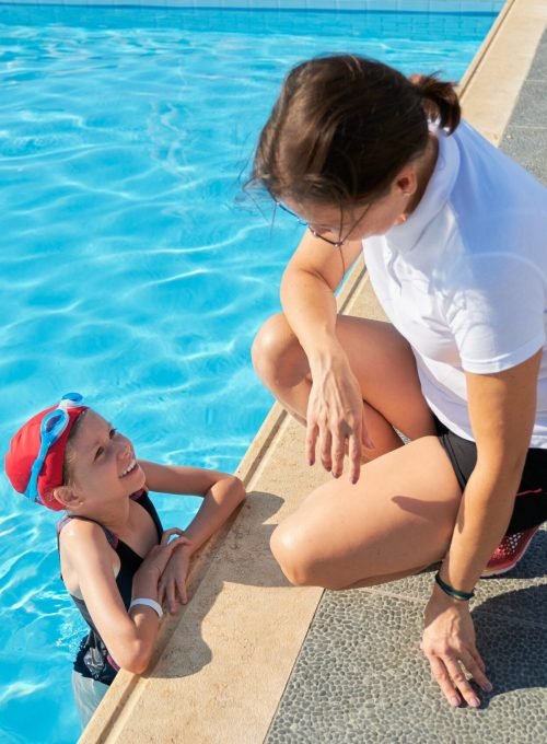 Active healthy lifestyle in children. Woman trainer talking to girl child in sports swimsuit hat and goggles for swimming near outdoor pool